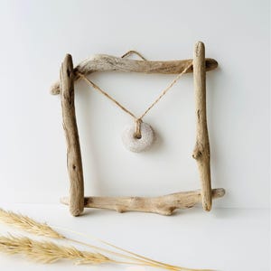 May include: A rustic driftwood frame with a natural stone pendant hanging in the center. The frame is constructed from weathered wooden branches, and the pendant is suspended by a twine cord. Dried wheat stalks are in the foreground.