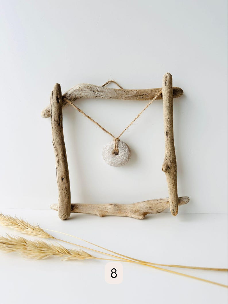 May include: A rustic driftwood frame with a natural stone pendant hanging in the center. The frame is constructed from weathered wooden branches, and the pendant is suspended by a twine cord. Dried wheat stalks are in the foreground.