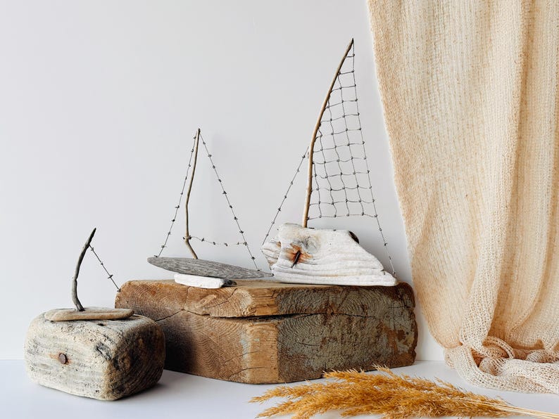 Puede incluir: Tres esculturas decorativas de veleros de madera. Los barcos est&aacute;n hechos de madera flotante y peque&ntilde;os bloques de madera. Las velas est&aacute;n hechas de malla de alambre y ramitas. Los barcos se exhiben sobre un bloque de madera y una superficie blanca.