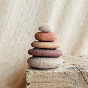 May include: A stack of five smooth, rounded stones arranged in a vertical pyramid shape. The stones vary in color, featuring shades of light gray, terracotta orange, tan, deep reddish-brown, and a speckled light gray base stone.