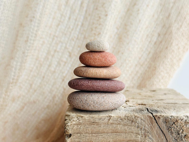 May include: A stack of five smooth, rounded stones arranged in a vertical pyramid shape. The stones vary in color, featuring shades of light gray, terracotta orange, tan, deep reddish-brown, and a speckled light gray base stone.