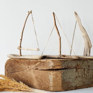 Puede incluir: Una escultura r&uacute;stica de madera con tres dise&ntilde;os abstractos de veleros hechos de madera flotante y ramitas. Los veleros descansan sobre un gran bloque de madera desgastada, con hierbas secas en primer plano. Los tonos naturales crean un tema costero y n&aacute;utico.