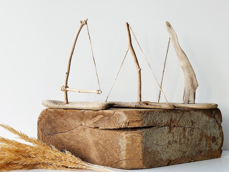 Puede incluir: Una escultura r&uacute;stica de madera con tres dise&ntilde;os abstractos de veleros hechos de madera flotante y ramitas. Los veleros descansan sobre un gran bloque de madera desgastada, con hierbas secas en primer plano. Los tonos naturales crean un tema costero y n&aacute;utico.