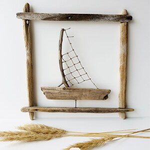May include: A handcrafted driftwood sailboat art piece. The boat features a wooden hull, a twig mast, and a net sail, set within a driftwood frame. The artwork is accented with dried grasses. The number 7 is visible in the lower left corner.