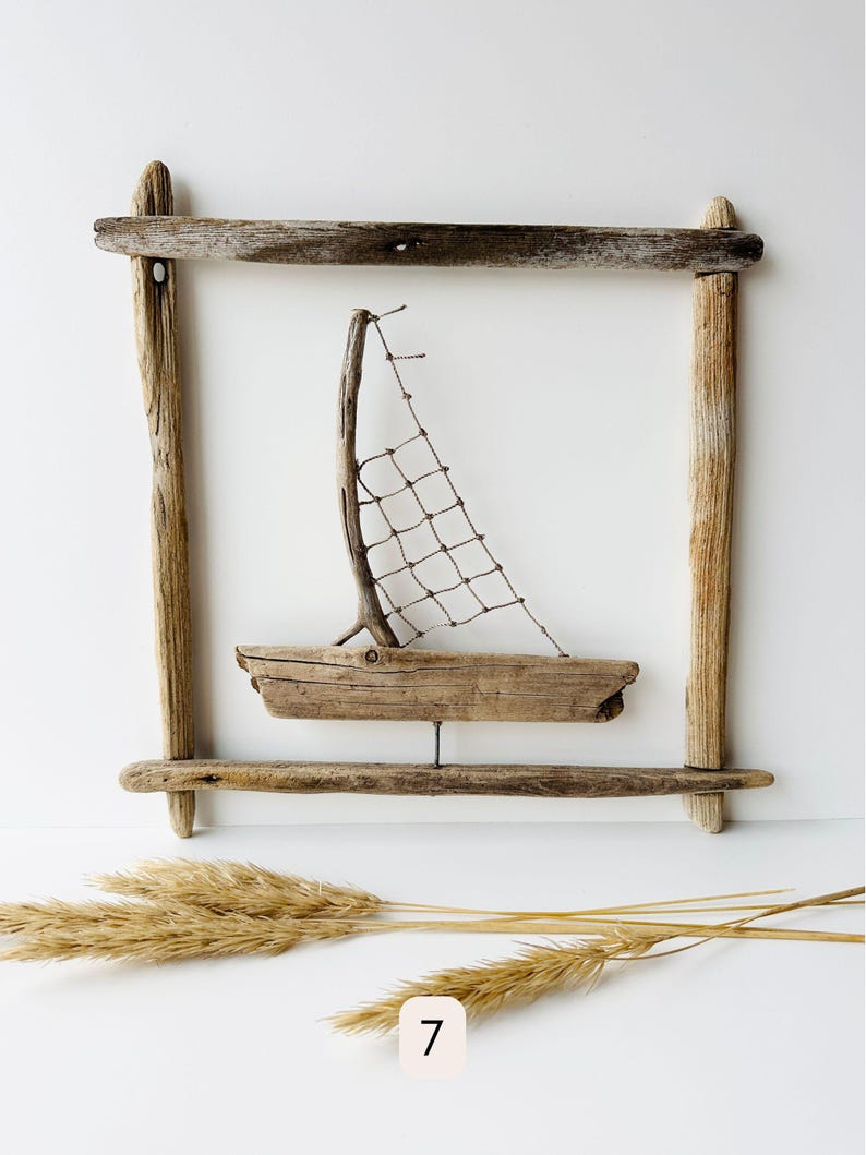 May include: A handcrafted driftwood sailboat art piece. The boat features a wooden hull, a twig mast, and a net sail, set within a driftwood frame. The artwork is accented with dried grasses. The number 7 is visible in the lower left corner.