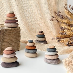 May include: Four small stacks of smooth, rounded river stones in various earth tones including shades of gray, tan, brown, and terracotta. Each stack is arranged vertically with the largest stones at the base and smaller stones at the top.