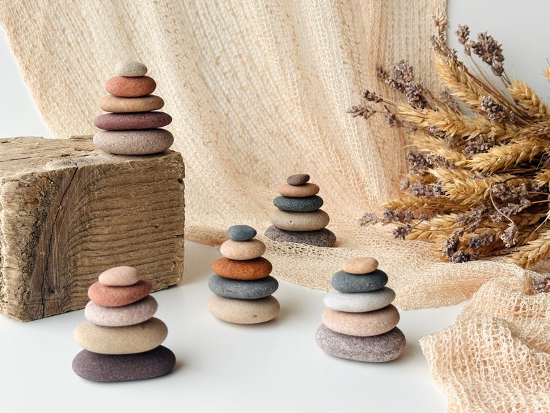 May include: Four small stacks of smooth, rounded river stones in various earth tones including shades of gray, tan, brown, and terracotta. Each stack is arranged vertically with the largest stones at the base and smaller stones at the top.