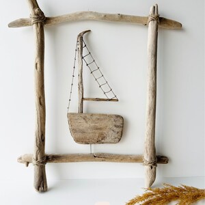 May include: A rustic wooden sailboat sculpture within a driftwood frame. The sailboat features a small sail made of netting and a wooden hull. The frame is constructed from weathered wood, tied together with twine, and has a natural, light brown color.
