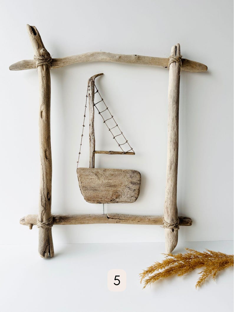 May include: A rustic wooden sailboat sculpture within a driftwood frame. The sailboat features a small sail made of netting and a wooden hull. The frame is constructed from weathered wood, tied together with twine, and has a natural, light brown color.