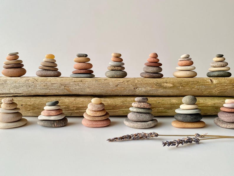May include: A collection of thirteen small stone cairns arranged in two rows on top of two pieces of weathered driftwood. The stones vary in color from tan, gray, and brown to soft pink and orange. Two sprigs of dried lavender lie in the foreground.