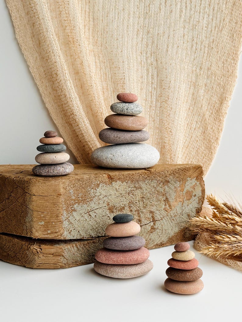 May include: Four stacks of smooth, rounded river stones in varying sizes and earth tones, including shades of gray, tan, pink, and brown. The stones are arranged in vertical columns, with the largest stack featuring five stones and the others four.