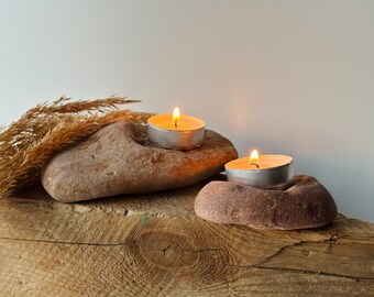 May include: Two lit tea light candles on natural stone candle holders, resting on a rustic wooden surface with dried grass.