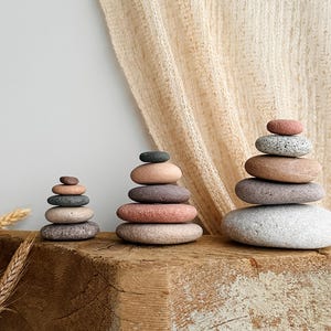 May include: Three stacks of smooth, rounded river stones arranged in ascending size order. The stones feature a natural color palette including shades of gray, tan, pink, and brown, with varying textures and speckled patterns on each individual rock.
