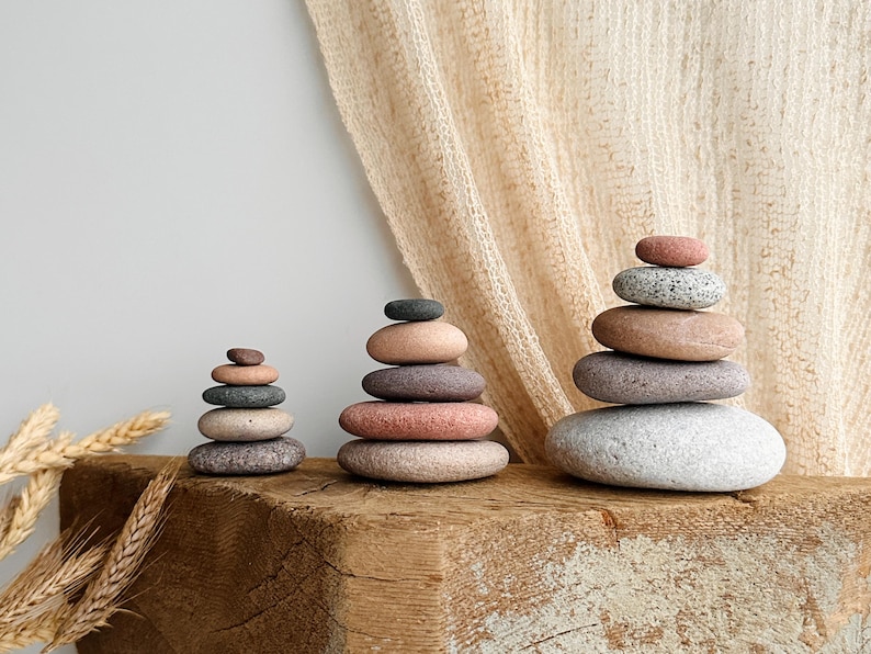 May include: Three stacks of smooth, rounded river stones arranged in ascending size order. The stones feature a natural color palette including shades of gray, tan, pink, and brown, with varying textures and speckled patterns on each individual rock.
