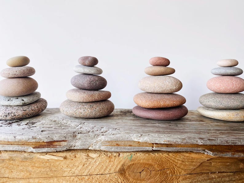 May include: Four separate stacks of smooth, rounded river stones arranged in vertical columns. Each stack consists of five or six stones of varying sizes, shapes, and natural earth tones, including shades of gray, tan, brown, and soft pink.