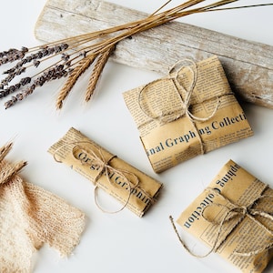 May include: Three rectangular packages wrapped in brown paper printed with vintage newspaper text and the words Original Graphing Collection, each tied with thin jute twine, accompanied by dried lavender and wheat stalks.