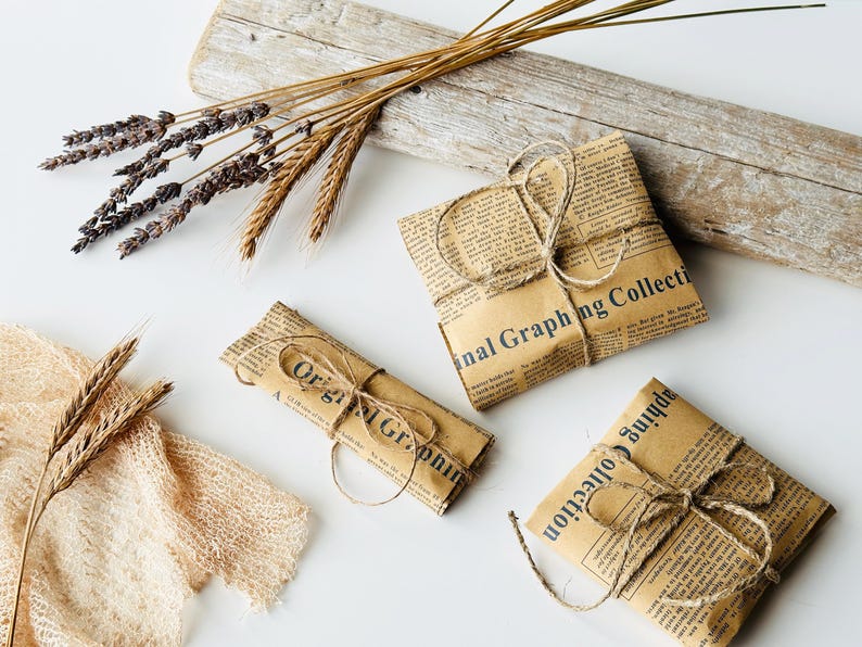 May include: Three rectangular packages wrapped in brown paper printed with vintage newspaper text and the words Original Graphing Collection, each tied with thin jute twine, accompanied by dried lavender and wheat stalks.