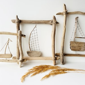 May include: Driftwood sailboat art pieces. Three wooden frames with small wooden sailboats inside. The sailboats have small sails and are made of natural wood. A bundle of dried wheat is in the foreground.