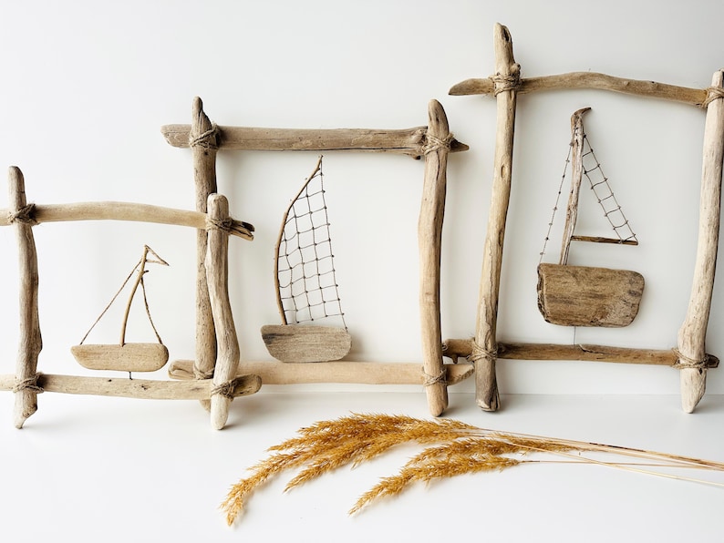 May include: Driftwood sailboat art pieces. Three wooden frames with small wooden sailboats inside. The sailboats have small sails and are made of natural wood. A bundle of dried wheat is in the foreground.
