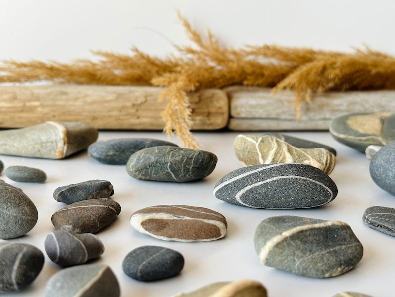 May include: An assortment of smooth, gray and brown river stones with white stripes and patterns. Dried grasses and weathered wood pieces are in the background, creating a natural, earthy aesthetic. The stones vary in size and shape.