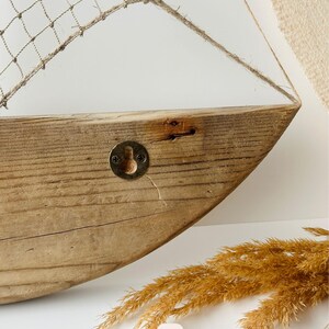 May include: A rustic wooden boat-shaped wall shelf with a net sail. The shelf is made of light brown wood with visible grain and a metal hook. The sail is made of a beige net and rope.