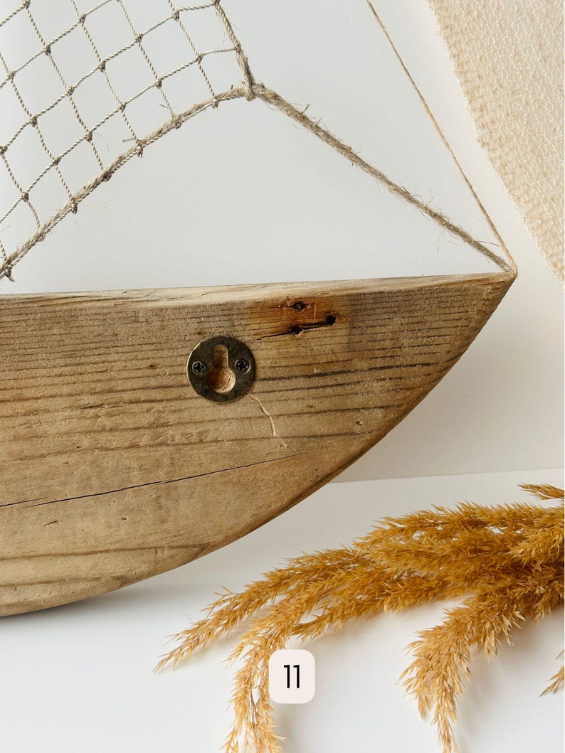 May include: A rustic wooden boat-shaped wall shelf with a net sail. The shelf is made of light brown wood with visible grain and a metal hook. The sail is made of a beige net and rope.