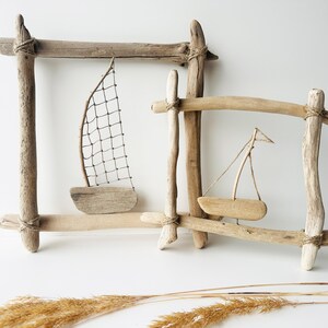 May include: Two driftwood frames with miniature wooden sailboats. One frame has a net sail, the other a triangular sail. The frames are constructed from natural wood pieces, tied together with twine, and set against a white background.