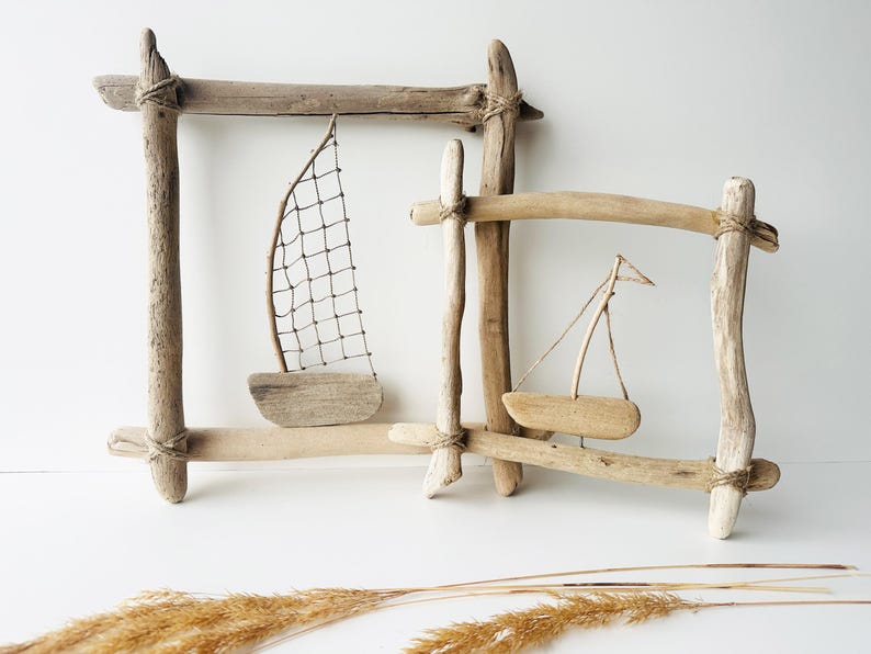 May include: Two driftwood frames with miniature wooden sailboats. One frame has a net sail, the other a triangular sail. The frames are constructed from natural wood pieces, tied together with twine, and set against a white background.