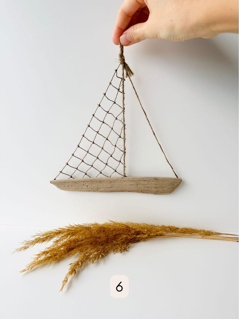 May include: A handcrafted sailboat decoration made from natural materials. The boat features a wooden hull, a net sail, and twine rigging. The image also includes a bundle of dried, golden-brown ornamental grass, and the number 6.