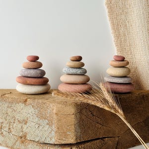 May include: Three stacks of smooth, rounded river stones in varying shades of terracotta, beige, gray, and cream. Each stack consists of five stones arranged in a pyramid shape, displaying natural textures and earthy color gradients.