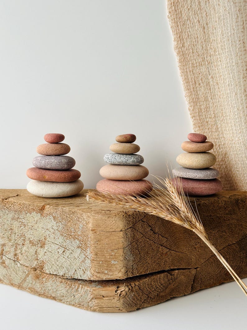 May include: Three stacks of smooth, rounded river stones in varying shades of terracotta, beige, gray, and cream. Each stack consists of five stones arranged in a pyramid shape, displaying natural textures and earthy color gradients.