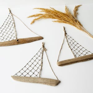 May include: Three driftwood sailboat wall hangings with netting sails and twine rigging. The boats are made of natural wood and are arranged on a white background. Dried wheat stalks are in the upper right corner.