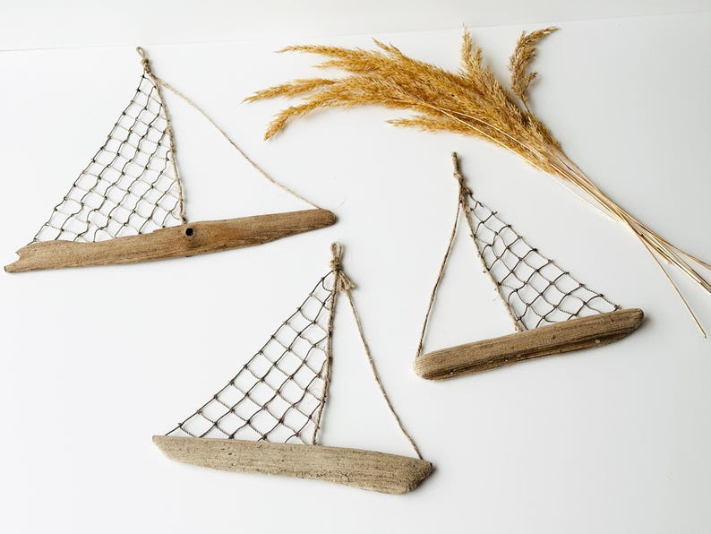 May include: Three driftwood sailboat wall hangings with netting sails and twine rigging. The boats are made of natural wood and are arranged on a white background. Dried wheat stalks are in the upper right corner.