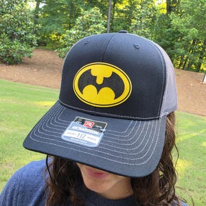 May include: A black and grey trucker hat featuring a yellow Batman logo on the front panel. The cap has a flat brim with grey stitching and a rectangular sticker that reads Richardson 112 Trucker Adjustable. The back panels are made of mesh.