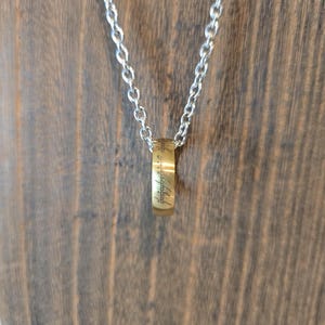 May include: A silver chain necklace with a gold-coloured ring pendant. The ring has engraved text. The necklace is displayed against a wooden background.