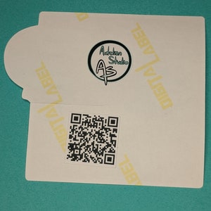 Może przedstawiać: Biała naklejka z czarno-białym kodem QR i czarnym k&oacute;łkiem z tekstem "Adobkin Studio AS" w kolorze białym. Naklejka ma tekst "DIGITAL LABEL" powt&oacute;rzony na ż&oacute;łto u g&oacute;ry i po prawej stronie.