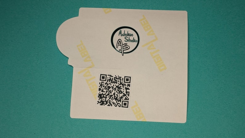 Może przedstawiać: Biała naklejka z czarno-białym kodem QR i czarnym k&oacute;łkiem z tekstem "Adobkin Studio AS" w kolorze białym. Naklejka ma tekst "DIGITAL LABEL" powt&oacute;rzony na ż&oacute;łto u g&oacute;ry i po prawej stronie.