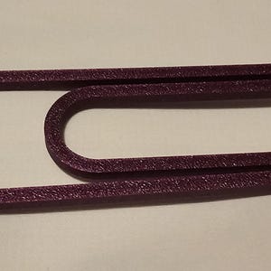 May include: A large, textured, purple paperclip. The metal clip is bent into a classic paperclip shape, with a rounded end and a straight section. The clip is a deep purple color.