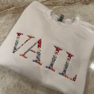 May include: White sweatshirt with the embroidered word "VAIL" in colorful floral letters. The letters are filled with red, orange, blue, and purple flowers and green leaves. The sweatshirt is folded on a marble surface.