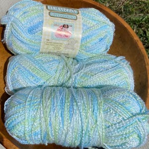 May include: Three skeins of Bernat Baby Coordinates yarn in a wooden bowl. The yarn is a mix of light blue, green, and white, with a metallic sheen. The label features a photo of a baby and a teddy bear.