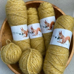 May include: Mustard yellow yarn skeins and balls are arranged in a wooden bowl. Each skein has a white label with a squirrel knitting. The labels read "Squirrel Wire" and "Net: 75g". The yarn is a warm, golden color.