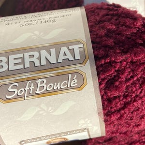 May include: A close-up of a skein of Bernat Soft Bouclé yarn in a deep burgundy color. The label indicates the yarn is 5 oz. / 140g. The yarn has a soft, textured appearance, ideal for knitting and crochet projects.