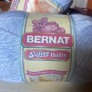 May include: A skein of Bernat Softee Baby yarn in light blue. The label features a pink teddy bear illustration and the text "Free Pattern on Label". Ideal for baby projects.
