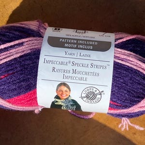 May include: A ball of Impeccable Speckle Stripes yarn in shades of purple, pink, and red. The label reads "Pattern Included" and "100% Acrylic". The yarn is suitable for knitting and crochet projects.