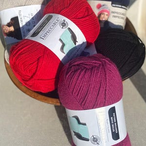 May include: A collection of yarn skeins in various colors, including red, black, and purple, are arranged in a wooden bowl. Each skein is labeled with the brand name "Impeccable" and includes details about the yarn's length and weight.