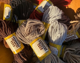 May include: A collection of colorful tapestry yarn skeins in shades of brown, red, gray, and black. Each skein has a yellow label with the text "TAPESTRY YARN" and "BUS CRAFTERS". The yarn is tightly wound and appears soft.