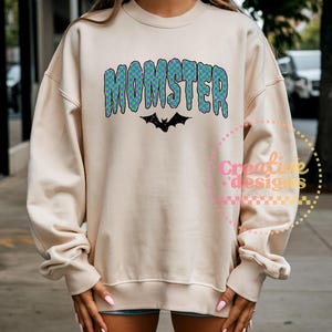 May include: A cream-colored sweatshirt with the word "MOMSTER" in blue and green checkered letters. A black bat graphic is below the word. The sweatshirt has long sleeves and a crew neck.