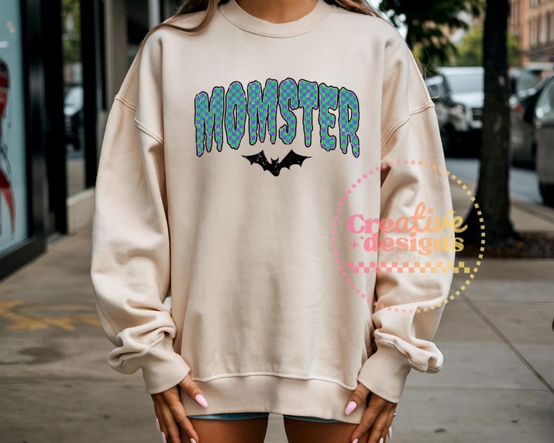 May include: A cream-colored sweatshirt with the word "MOMSTER" in blue and green checkered letters. A black bat graphic is below the word. The sweatshirt has long sleeves and a crew neck.