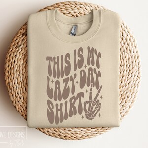 Puede incluir: Una sudadera beige con el texto "This is my lazy day shirt" en letras marrones onduladas. Una mano de esqueleto marr&oacute;n con un signo de paz est&aacute; a la derecha del texto.