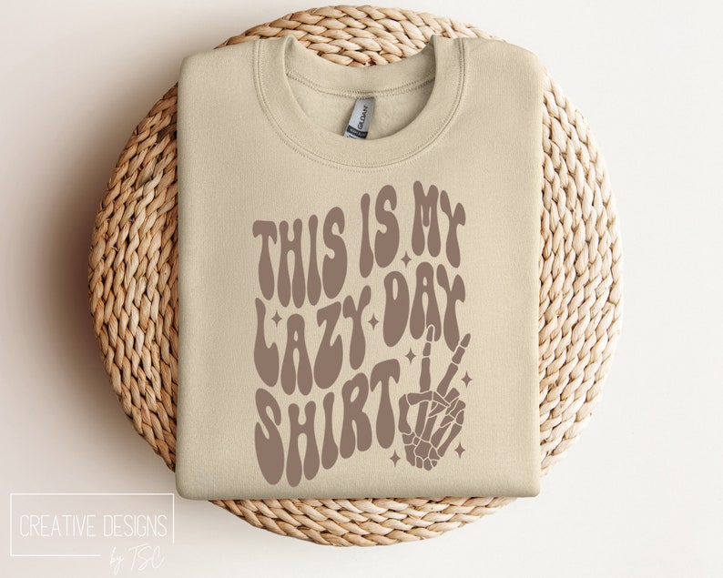 Puede incluir: Una sudadera beige con el texto "This is my lazy day shirt" en letras marrones onduladas. Una mano de esqueleto marr&oacute;n con un signo de paz est&aacute; a la derecha del texto.